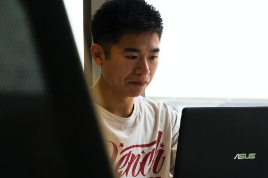 A young man with dark hair looks down at a laptop screen, partially obscured by a dark chair in the foreground. He is wearing a white t-shirt with red text.